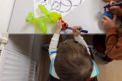 Dziecko w kolorowej kamizelce rysuje po kartce z konturami postaci, trzymając w ręku gruby, niebieski marker. Na stole przed nim leży zielony balon zwierzątko, którego kształt przypomina psa. Obok dziecka widać rękę innego uczestnika trzymającą pomarańczowy marker.
