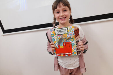 Dziewczynka z uśmiechem trzyma kolorową książkę z ilustracją lokomotywy. Jest ubrana w różową bluzę i spodnie, a na ramieniu ma zawieszoną różową torbę. Stoje przed białą ścianą i w tle widoczna jest część ekranu multimedialnego.