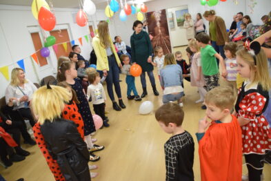 Grupa dzieci w kolorowych strojach bawi się w przestronnym, dobrze oświetlonym pomieszczeniu przyozdobionym balonami. Dorośli również są obecni, obserwując zabawy i uczestnicząc w interakcji z dziećmi. Ściany są udekorowane zdjęciami i plakatami związany z kolejnictwem.