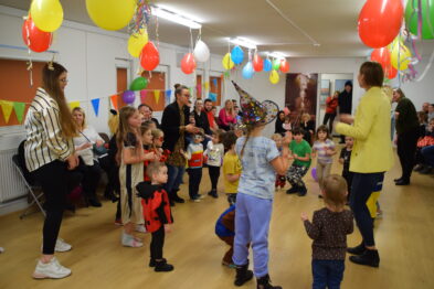 Grupa dzieci uczestniczy w zabawach w pomieszczeniu ozdobionym kolorowymi balonami i dekoracjami karnawałowymi. Dzieci, niektóre w przebraniach, są zaangażowane w grę pod kierunkiem dorosłych. W tle znajduje się stół z napojami i przekąskami dla uczestników.