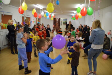 Grupa dzieci bawi się w przestronnym wnętrzu zdobionym kolorowymi balonami wiszącymi na suficie. Niektóre dzieci mają na sobie kolorowe kostiumy, a w rękach trzymają zabawki lub balony. Wokół dzieci dorosłe osoby obserwują zabawę lub uczestniczą w wydarzeniu, między innymi trzymając balony.