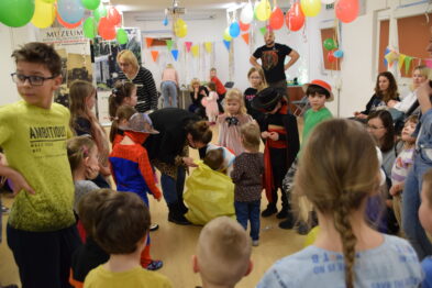 Grupa dzieci w kostiumach wraz z dorosłymi uczestniczy w uroczystości w pomieszczeniu zdobionym balonami i kolorowymi dekoracjami. Na pierwszym planie dziecko ma na sobie żółty strój, a inne dzieci otaczają kogoś w czarnej pelerynie i kapeluszu. Sala jest pełna i tętni aktywnością, a uczestnicy eventu zajęci są wspólną zabawą.