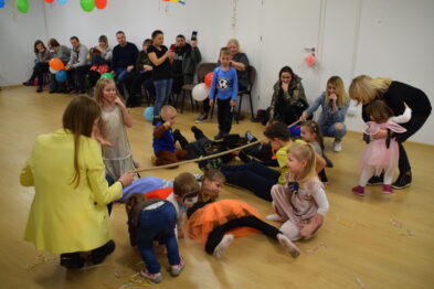 Dzieci uczestniczą w zabawie na podłodze w jasnym pomieszczeniu, w którym widoczna jest grupa dorosłych obserwatorów. W tle pomieszczenia widać balony oraz siedzących dorosłych. Dzieci i opiekunowie są ubrani w codzienne stroje.
