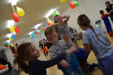 Dzieci bawią się w wypełnionym kolorowymi balonami wnętrzu, chwytając pęcherzyki mydlane unoszące się w powietrzu. Uśmiechnięta dziewczynka w pierwszym planie wydaje się być rozbawiona, patrząc na unoszące się przed nią pęcherzyki. W tle inni uczestnicy imprezy tańczą i bawią się, tworząc radosną atmosferę karnawałowej zabawy.