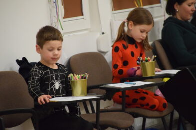 Dzieci w kostiumach siedzą przy stolikach i zajmują się rysowaniem. Chłopiec po lewej ma na sobie czarny strój z motywem pająka, a dziewczynka po prawej jest przebrana za biedronkę. W tle widoczna jest dorosła osoba obserwująca ich zajęcia.