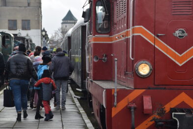Grupa ludzi w różnym wieku, w tym dzieci, wsiada do czerwonego pociągu o wyglądzie retro stojącego na torach. Peron jest mokry i wydaje się być chłodny dzień. W tle widoczne są historyczne zabudowania skansenu kolejowego.