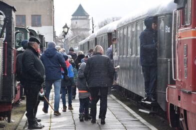 Grupa ludzi stoi na peronie obok zabytkowych, wąskotorowych wagonów kolejowych. Passażerowie w różnym wieku ubrani w ciepłe kurtki wsiadają do pociągu lub rozmawiają między sobą na dworze. Na tle zimowej aury, w tle widać historyczne budynki kolejowe.
