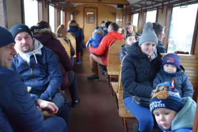 W drewnianym wnętrzu wagonu wąskotorowego pociągu siedzą ludzie na ławkach ustawionych naprzeciwko siebie. Dzieci i dorośli są ubrani w ciepłe zimowe ubrania, kilka osób uśmiecha się, widać rozmowy, jeden z chłopców nosi czapkę z napisem. Przez okna wagonu można dostrzec widok na zewnątrz, lecz szczegóły są niewyraźne.