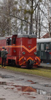 Czerwona lokomotywa wąskotorowa stoi na torach z przyczepionymi do niej wagonami pasażerskimi, podczas gdy ludzie rozchodzą się po peronie. Grupa osób jest ubrana w różnego rodzaju odzież, odzież odblaskową oraz kolorowe kurtki. Wilgotne tory i peron odzwierciedlają pochmurne niebo, a na ziemi widać kałuże.