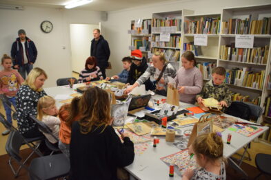 Grupa dzieci i dorosłych zajmuje się twórczymi aktywnościami przy stole w wypełnionym książkami pomieszczeniu, które wygląda na bibliotekę lub czytelnię. Na stole rozłożone są artykuły plastyczne, takie jak papier, farby i kredki, a uczestnicy tworzą różnorodne prace rękodzielnicze. Wokół stołu zgromadziły się osoby w różnym wieku, intensywnie zaangażowane w wykonywanie swoich projektów.