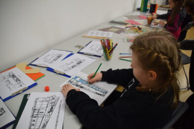 Dzieci siedzą przy długim stole i zajmują się kolorowaniem oraz rysowaniem. Na stole rozłożony jest różnorodny materiał plastyczny, tak jak kredki, flamastry i arkusze papieru z konturami pociągów. Przy jednym z końców stołu widać stojące kubki z przyborami do pisania.