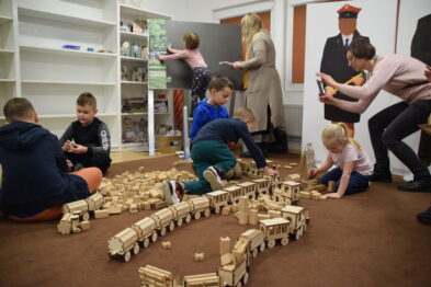 Dzieci siedzą na podłodze i składają drewniane modele kolejowe. W tle widać dorosłych, którzy nadzorują i pomagają młodszym uczestnikom. Pomieszczenie jest jasne i wydaje się być obszarem wyznaczonym na zajęcia grupowe lub warsztaty.