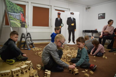Grupa dzieci siedzi na podłodze i bawi się drewnianymi klockami, tworząc konstrukcje przypominające elementy kolejowe. W tle dorosłych i inne dzieci oglądają wystawę lub również uczestniczą w zabawie. Pomieszczenie jest jasne, a na ścianie widać plakat z motywem kolejowym.