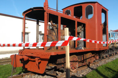 Lokomotywa spalinowa o czerwonej kabine z elementami w kolorze czarnym stoi na torach kolejowych. Wokół lokomotywy ustawiono taśmę ostrzegawczą w biało-czerwone pasy. Maszyna posiada widoczne wyposażenie, takie jak bufor czy sprzęg kolejowy.