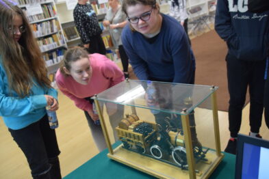 Grupa uczniów ogląda model lokomotywy znajdujący się w szklanej gablocie umieszczonej na podwyższeniu zabezpieczającym eksponat. Dzieci wydają się zaciekawione eksponatem, badają go wzrokiem. W tle widoczne są kolejne elementy ekspozycji muzealnej i fragmenty książek na półkach, co sugeruje, że scena ma miejsce w bibliotece lub czytelni.