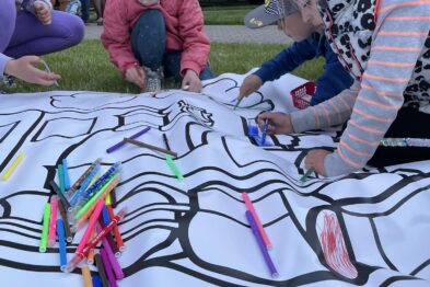 Dzieci klęczą na trawie i wspólnie malują na dużym arkuszu papieru z wydrukowanymi torami kolejowymi. Na arkuszu leży wiele kolorowych flamastrów, które maluchy używają do dekorowania rysunku. W tle można dostrzec fragmenty zielonego krajobrazu i elementy zabytkowego taboru kolejowego.