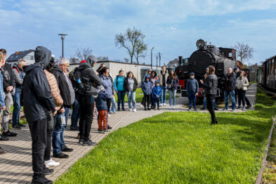 Grupa osób zebrała się na zewnątrz wokół przewodnika, który opowiada im o atrakcjach muzeum. W tle widać klasyczną, czarną lokomotywę parową wąskotorową zwaną również parowozem. Na pierwszym planie można dostrzec torowisko, które wykorzystywane jest do prezentacji pojazdów kolejowych.