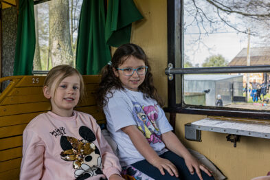 Dwie młode dziewczynki siedzą na drewnianej ławce wewnątrz wagonu kolejowego, jedna z nich ma na sobie bluzę z postacią kreskówkową. Obok otwartego okna, przez które widać zewnątrz budynek i przybyłych gości, zasłony są odgarnięte i zwisają w kolorze zielonym. Obie pasażerki zdają się być zadowolone, patrząc w kierunku kamery.
