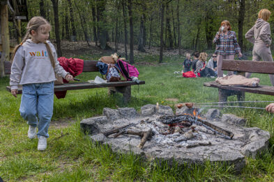 Dzieci bawią się na zewnątrz w pobliżu ogniska, na którym piecze się kiełbaski. W tle widać kolej wąskotorową i kilka osób siedzących na ławce. W otoczeniu drzew i zieleni, atmosfera wydaje się luźna i piknikowa.