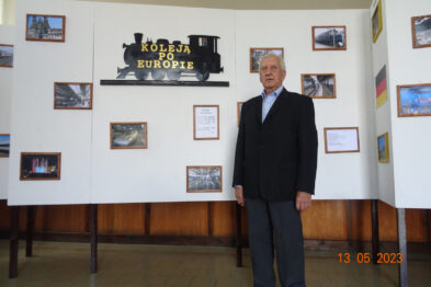 Starszy mężczyzna w ciemnym garniturze stoi wewnątrz pomieszczenia z wystawą dotyczącą kolei. Za nim znajduje się ściana z wieloma kolorowymi obrazkami i plakatami, a nad nimi wisząca czarna tablica z białym pociągiem i napisem 