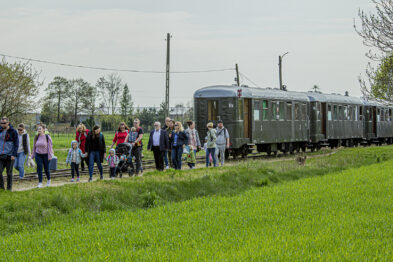 Grupa osób stoi na zielonej łące w pobliżu torów kolejowych, gdzie zatrzymał się stary wagon kolejowy. Osoby są skierowane w stronę fotografa, niektóre machają do kamery. W tle widać niebieskie, częściowo zachmurzone niebo oraz elementy infrastruktury kolejowej.