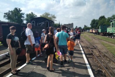 Grupa osób czeka na peronie obok torów kolejowych w towarzystwie parowozu o czarnym kolorze. Niektórzy z uczestników rozmawiają, inni patrzą w stronę aparatu fotograficznego. Słońce świeci, a na niebie widoczne są nieliczne chmury.