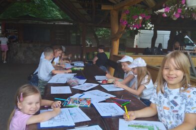 Grupa dzieci siedzi przy długim drewnianym stole pod zadaszoną wiatą, zajmując się kolorowaniem i pisaniem. Uśmiechnięte dzieci są skupione na kartach leżących przed nimi, używając różnych kolorowych kredek. W tle widać zieloną roślinność i innych uczestników imprezy w rozmytym planie.