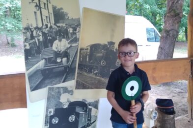Chłopiec w okularach stoi przed roll-upem promującym Muzeum Kolei Wąskotorowej; dzierży w rękach drewnianą tarczę z zielonym sygnałem kolejowym. Z tyłu widoczne są elementy drewnianej konstrukcji, prawdopodobnie peronu lub wiaty. W tle plakat z czarno-białymi zdjęciami przedstawiającymi pociąg i tory kolejowe wskazuje na kolejowy charakter wydarzenia.