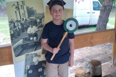 Chłopiec stoi uśmiechnięty, trzymając w ręce zielono-białą chorągiewkę kolejową, obok plakatu promującego 