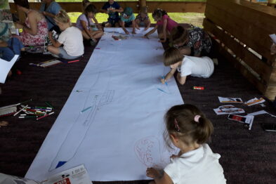Grupa dzieci siedzi na podłodze i rysuje kredkami na długim arkuszu papieru rozłożonym przed nimi. Wokół rozrzucone są kolorowe kredki i kawałki papieru. Dzieci są skupione na swojej twórczości, a tło tworzą drewniane ławki i fragmenty konstrukcji, w której się znajdują.