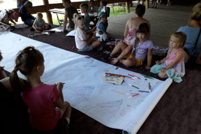 Grupa dzieci siedzi na podłodze i rysuje kolorowymi kredkami na dużym arkuszu papieru rozłożonym przed nimi. Są skupione na tworzeniu rysunków, a przestrzeń, w której przebywają, wygląda na zadaszony i otwarty obszar. Nieopodal dzieci widać kosz pełen kredek i pisaków, które są wykorzystywane podczas tej kreatywnej aktywności.