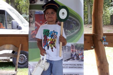 Małe dziecko stoi na drewnianym podestie trzymając zieloną deskę z wyraźnym numerem oraz biały megafon. Jest ubrane w białą koszulkę z kolorowym nadrukiem, czapkę i czerwono-niebieskie buty. W tle widać roll-up z napisem 
