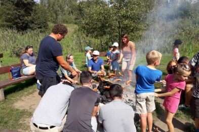 Grupa ludzi, w tym dorosłych i dzieci, gromadzi się wokół ogniska, na którym gotowane jest jedzenie. Otoczeni są zielenią i siedzą na ławkach bądź stoją na trawie, a nad płomieniami unosi się dym. Widać, że uczestnicy są zaangażowani w wspólne przygotowywanie posiłku na świeżym powietrzu.