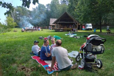 Grupa osób siedzi na kocach rozłożonych na trawie w parku, skupiając uwagę na wspólnym pikniku. W tle widoczna jest drewniana altanka i drzewa, a w powietrzu unosi się dym z przygotowywanego posiłku na grillu. Na pierwszym planie znajduje się wózek dziecięcy, a wokół rozmieszczone są akcesoria piknikowe.