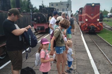 Grupa osób czeka na peronie kolejowym; w tle widać zieloną lokomotywę z czerwonym przodem. Dorośli i dzieci są ubrani w letnie stroje; jedno z dzieci ma na głowie różowy kapelusz. Osoby stoją w kolejce obok stojącego wagonu, a w tle widać budynek stacji kolejowej.