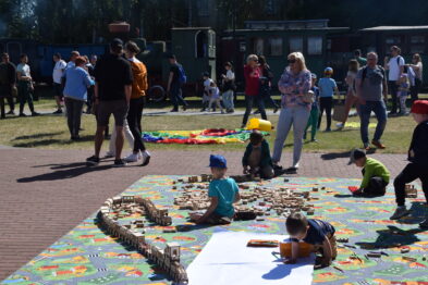 Dzieci bawią się na kolorowej macie w plenerze, układając tor kolejowy i rysując. W tle widać grupy ludzi stojących oraz spacerujących w słoneczny dzień. Wokół miejsca zabaw rozmieszczone są zabawki i miękka mata, tworząc przestrzeń dla kreatywnej zabawy.