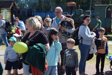 Grupa ludzi, w tym dzieci i dorośli, stoi na zewnątrz w słoneczny dzień. Niektóre z dzieci trzymają balony, a w tle widoczne są zielone drzewa i wagon kolejowy. Osoby na zdjęciu wydają się być skupione na wydarzeniu, które obserwują, i noszą różnorodne, luźne ubrania.