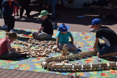 Dzieci w różnokolorowych czapkach siedzą na ziemi i bawią się drewnianymi klockami, układając z nich konstrukcję przypominającą tor kolejowy i pociąg. Do zabawy używają dużych, kolorowych klocków. Miejsce zabawy to duża, jasna przestrzeń na zewnątrz, pokryta kolorowymi matami.