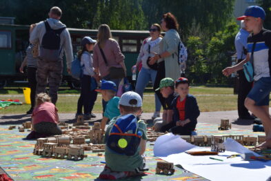 Dzieci w różnym wieku bawią się klockami na dużym, kolorowym dywanie na zewnątrz, składając z nich modele kolejowe. Osoby dorosłe stoją i siedzą wokół, obserwując młodych konstruktorów. W tle widoczne są zielone drzewa i fragment wagonu kolejowego.