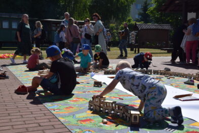Dzieci bawią się na zewnątrz drewnianymi modelami kolejek na kolorowej, dużej makiecie z torami. Kilka osób w tle obserwuje zabawę, a dzieci koncentrują się na układaniu i przesuwaniu pociągów. Całość odbywa się na słonecznym dziedzińcu z ceglaną nawierzchnią.
