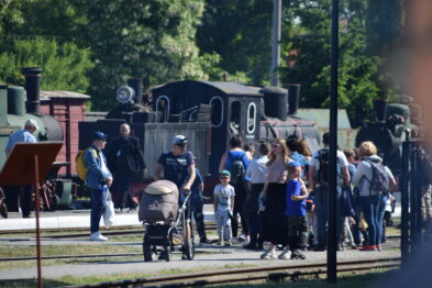 Na zdjęciu widoczni są ludzie, w tym dorośli i dzieci, zgromadzeni na terenie muzealnym wśród eksponatów kolejowych. W tle dostrzegalna jest stara parowa lokomotywa, która stanowi atrakcję dla odwiedzających. Wskazuje to na aktywne uczestnictwo publiczności w tematycznym evencie związanym z kolejnictwem.