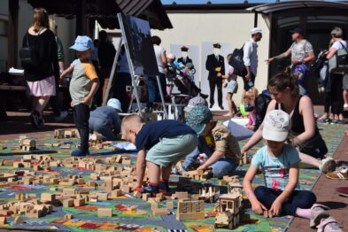 Dzieci w kapeluszach i daszkach bawią się drewnianymi klockami, układając je na ziemi, tworząc kolorową makietę miasta lub torów kolejowych. Dorośli i inni obserwatorzy otaczają miejsce zabawy, a na tle widać zabudowania, które mogą być częścią muzealnego kompleksu. Słońce świeci, co wskazuje na pogodny dzień, podczas gdy uczestnicy eventu są skupieni na wspólnej zabawie.