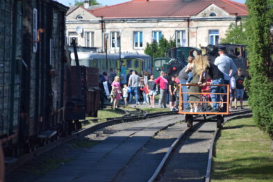 Grupa osób, w tym dzieci, jest widoczna na torach kolejowych, gdzie niektórzy z nich jadą małym wagonikiem kolejowym. W tle widać budynek, który może być stacją kolejową oraz zaparkowane kolejne wagony. Otoczenie jest słoneczne i uporządkowane, co dodaje uroku spotkaniu w muzealnym otoczeniu.