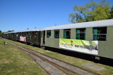 Na szynach stoi zielony wagon kolejowy, do którego przyczepiony jest duży, kolorowy baner informujący o 