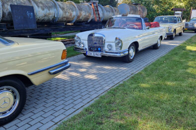 Klasyczne samochody marki Mercedes zaparkowane są w rzędzie na terenie muzeum kolejnictwa, z wagonami kolejowymi w tle. Na pierwszym planie widoczny jest kremowy kabriolet z otwartym dachem i chromowanymi detalami. Po lewej stronie zdjęcia częściowo widać inny samochód w kolorze żółtym.