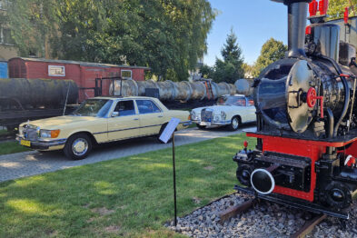 Stojący na trawiastym podłożu, żółty mercedes obok czarnej lokomotywy parowej jest eksponowany na terenie muzeum. Pojazd ma otwarte drzwi kierowcy, a przed nim umieszczono tabliczkę informacyjną. W tle widać inne klasyczne samochody oraz elementy infrastruktury kolejowej.