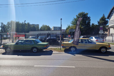 Dwa klasyczne samochody marki Mercedes, jeden zielony i drugi żółty, stoją obok siebie na asfaltowym placu. Między pojazdami umieszczono reklamowy baner z logo MB/8 Club Poland. Tło stanowi budynek z białymi i szarymi elementami oraz metalowe ogrodzenie, za którym widać zarysy kolejowego taboru.