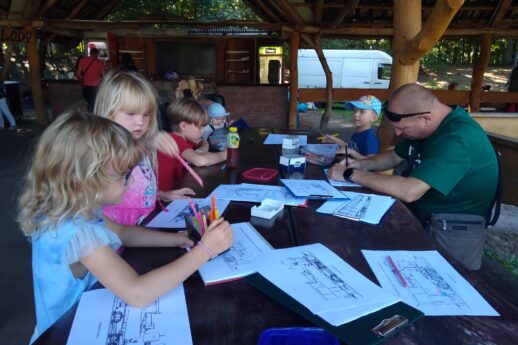 Dzieci siedzą przy drewnianych stołach i kolorują obrazki przedstawiające różne pojazdy szynowe. W tle widać zielone drzewa i część budynku, który wydaje się być altanką lub zadaszonym pawilonem. Dorosły mężczyzna wspomaga młodszych uczestników, towarzysząc im w ich kreatywnej aktywności.