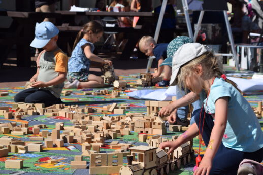 Dzieci bawią się drewnianymi klockami na otwartej przestrzeni, układając z nich makietę miasta i torów kolejowych. Na pierwszym planie widoczna jest dziewczynka skupiona na zabawie, a w tle inni uczestnicy również zaangażowani w tworzenie konstrukcji. Wokół dzieci rozłożone są liczne elementy zabawki, w tym kolorowe domki i fragmenty szyn.