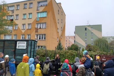 Grupa osób stoi na chodniku, słuchając prezentacji kobiety, która stoi przed nimi i trzyma coś w rękach. Uczestnicy, ubrani w kurtki i kolorowe czapki, pozornie uważnie słuchają. W otoczeniu są miejskie budynki mieszkalne i fragmenty zieleni.